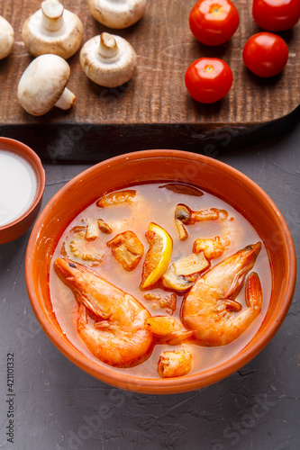 Tom yam soup with shrimps in a plate on a wooden table on a napkin next to a bowl with coconut milk mushrooms and tomatoes.