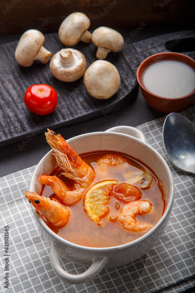 Tom yam soup with shrimps in a tureen on a concrete background next to a bowl with coconut milk tomatoes and mushrooms on a wooden stand. Tom yam soup with shrimps in a tureen on a concrete background next to a bowl with coconut milk tomatoes and mushrooms on a wooden stand.