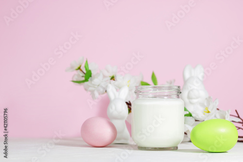 Organic probiotic milk kefir drink or yogurt in a glass container, with colored Easter eggs, on a pink background with a rabbit. Easter concept. Cold fermented milk drink with probiotics.