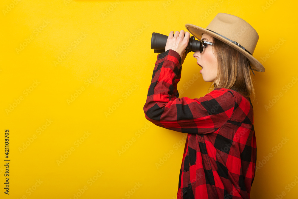 Young woman with a surprised face in a hat and a plaid shirt looks through binoculars on a yellow background