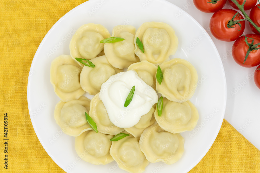 Boiled Russian dumplings with sour cream and tomatoes on a plate on a yellow napkin. Top view. Boiled Russian dumplings with sour cream and tomatoes on a plate on a yellow napkin. Top view.