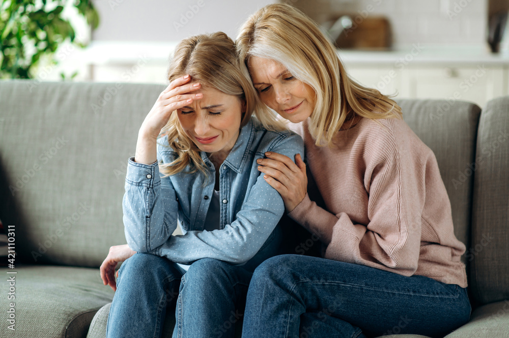 Mother and daughter together at living room. Loving mature mom sits on the couch, hugs her young adult crying daughter, stroking her, sympathizes, family relationship and values concept Mother and daughter together at living room. Loving mature mom sits on the couch, hugs her young adult crying daughter, stroking her, sympathizes, family relationship and values concept