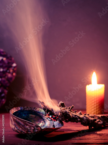 Smudging sage with dense smoke. Copy space. Amethyst crystal and burning bees wax candle at background. Smudging sage, amethyst and beeswax candles can be used to cleanse your home of negative energy.