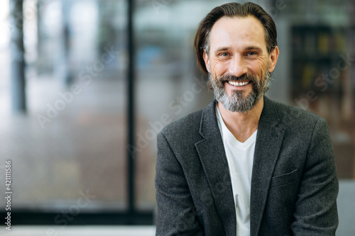 Portrait of a handsome office worker or ceo. Confident stylish bearded middle aged caucasian man in formal jacket and white t-shirt smiles and looks directly into the camera