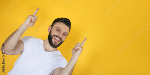 Excited handsome young bearded man in a white t-shirt is pointing away over yellow background