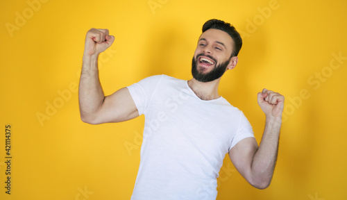Handsome excited modern young bearded stylish man is posing with a wide toothy smile on yellow background in studio