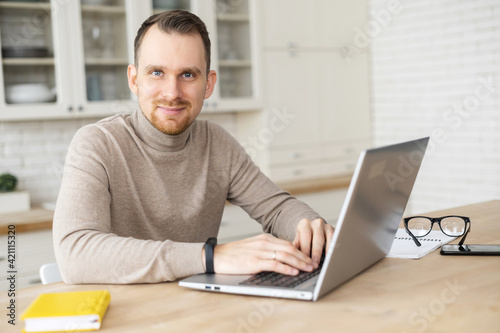 Portrait of friendly attractive male with a laptop at home, a blue-eyed bearded man looks at the camera, smiles. Remotely work concept