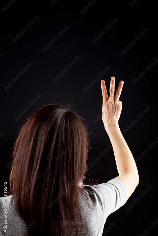 Behind the head view of a young caucasian woman with long brown hair wearing short sleeve shirt against dark background. She shows three fingers with her hand. A versatile concept image for number 3 Behind the head view of a young caucasian woman with long brown hair wearing short sleeve shirt against dark background. She shows three fingers with her hand. A versatile concept image for number 3