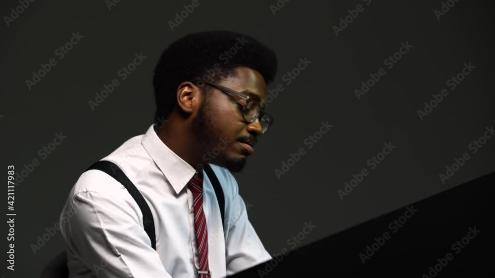 Handsome African American man in a stylish suit and glasses sings and composes himself on the grand piano with pleasure. A black performer performs on a gray background in the studio. Close up. Handsome African American man in a stylish suit and glasses sings and composes himself on the grand piano with pleasure. A black performer performs on a gray background in the studio. Close up.