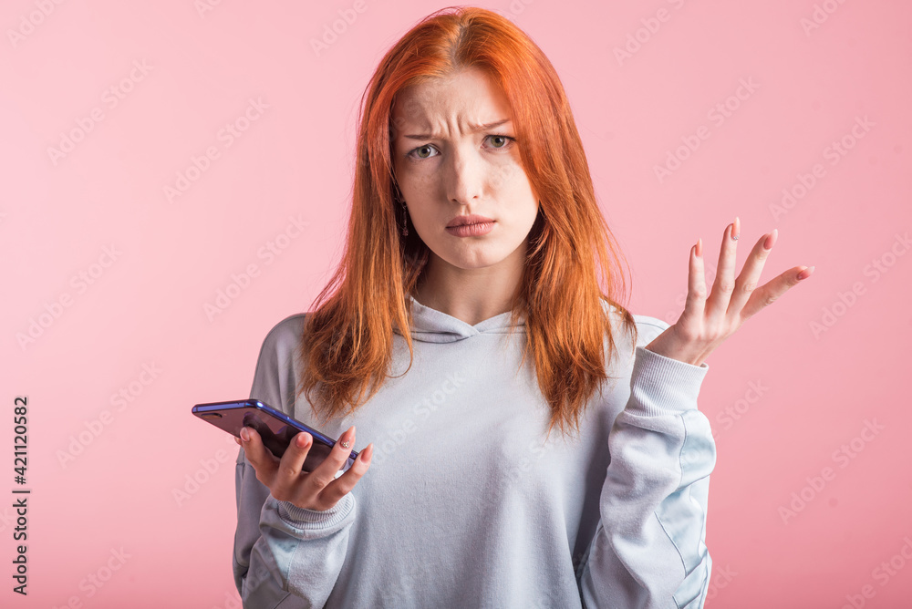 Confused redhead girl with phone in studio on pink background Confused redhead girl with phone in studio on pink background
