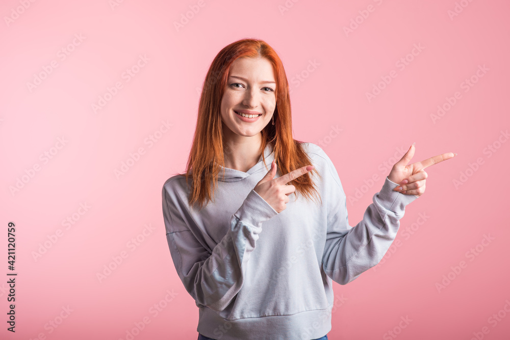 Redhead girl points index fingers to the side for copyspace in studio on pink background