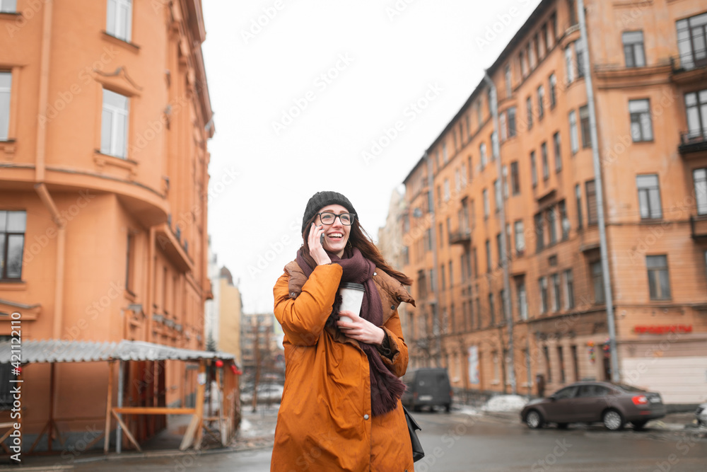 Happy young woman with glass of coffee in her hands, talking on phone on walk.