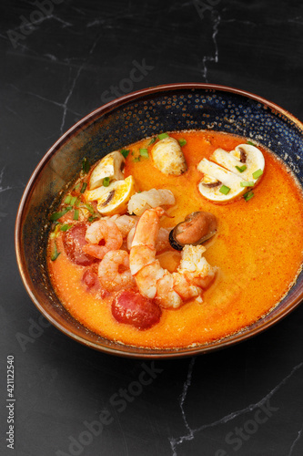 Asian soup Tom Yam with shrimps, mussels, mushrooms, tomatoes and spring onion. Dish isolated in a blue bowl, close-up on a black marble background. Asian cuisine.