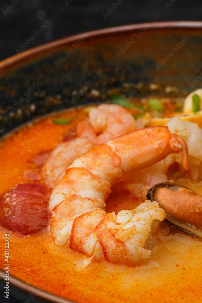 Asian soup Tom Yam with shrimps, mussels, mushrooms, tomatoes and spring onion. Dish isolated in a blue bowl, close-up on a black marble background. Asian cuisine. Asian soup Tom Yam with shrimps, mussels, mushrooms, tomatoes and spring onion. Dish isolated in a blue bowl, close-up on a black marble background. Asian cuisine.