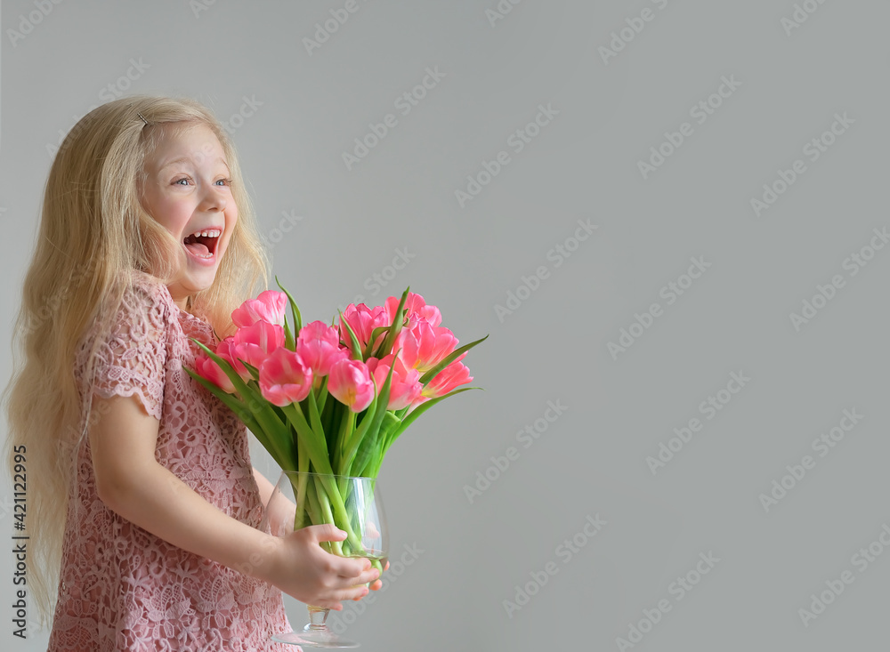 Laughing cute little blonde girl holding a vase of pink tulips on the grey background