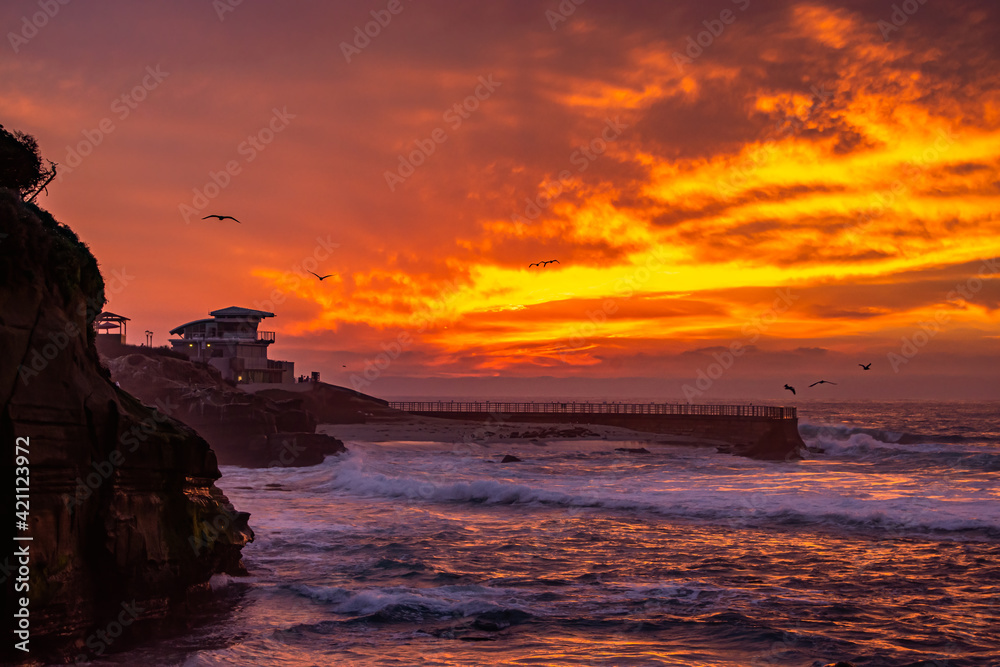 Dramatic fiery sunset over the waves crashing onto the beach with La Jolla Children's pool in background in San Diego California with seagulls flying in the air