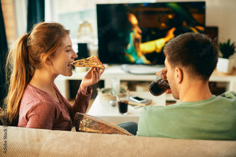 Happy woman eating pizza while watching movie with her boyfriend at home.