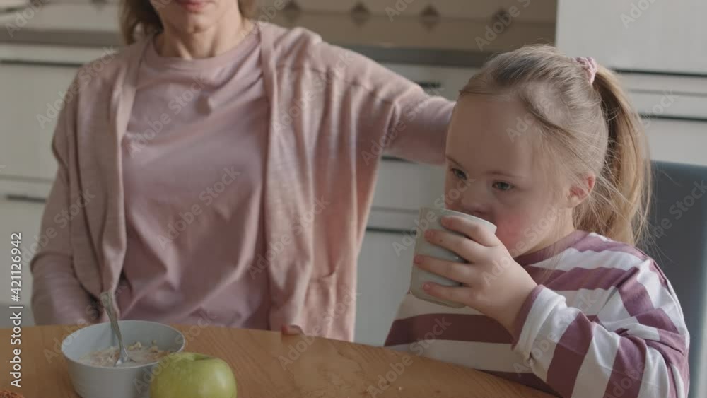 Medium shot of unrecognizable woman sitting at kitchen table and patting on head her lovely little daughter with down syndrome while she drinking juice during breakfast Medium shot of unrecognizable woman sitting at kitchen table and patting on head her lovely little daughter with down syndrome while she drinking juice during breakfast
