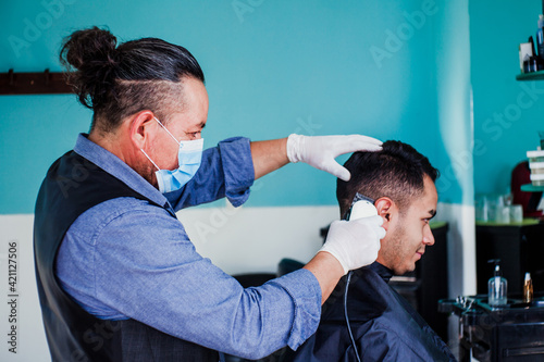 hispanic man stylist with facemask cutting hair to a client in a barber shop in Mexico city in coronavirus pandemic