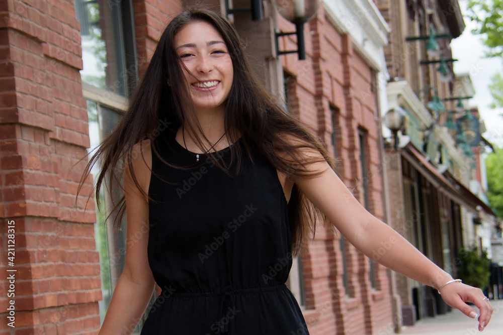 Outdoor fun portrait of beautiful young woman in sleeveless black dress smiling while spinning on the city sidewalk.
