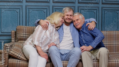 Family of senior grandparents with adult son embracing having fun looking at camera bonding at home
