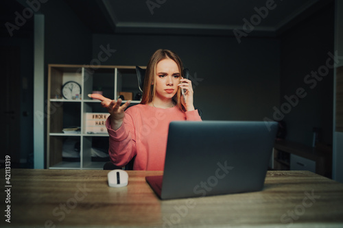 Business woman works at home at a table on a laptop and calls on a smartphone and looks at the computer screen. Beautiful woman working remotely from home on laptop.