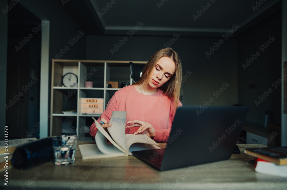 Obraz premium Female student in a pink sweater studies remotely at home, looks in a notebook with a serious face and uses a laptop. Home learning concept.