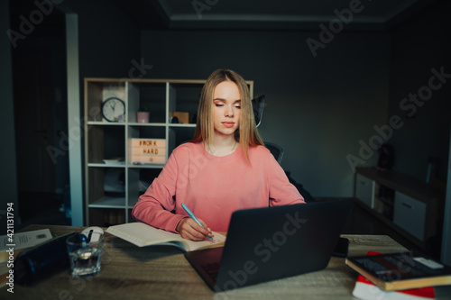 Beautiful student girl sitting at home at a desk with a laptop and studying online, looking at the screen and copying in a notebook