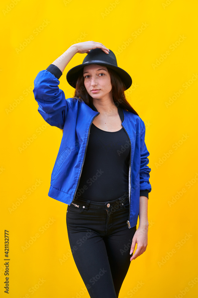 Vertical photo portrait of a young caucasian woman in black hat and blue jacket on yellow background, holding hat with hand Vertical photo portrait of a young caucasian woman in black hat and blue jacket on yellow background, holding hat with hand