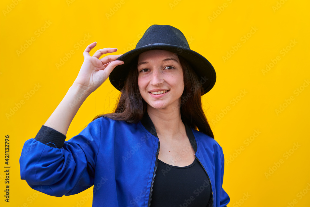 Portrait of a young caucasian woman in black hat and blue jacket on yellow background, holding hat with hand Portrait of a young caucasian woman in black hat and blue jacket on yellow background, holding hat with hand