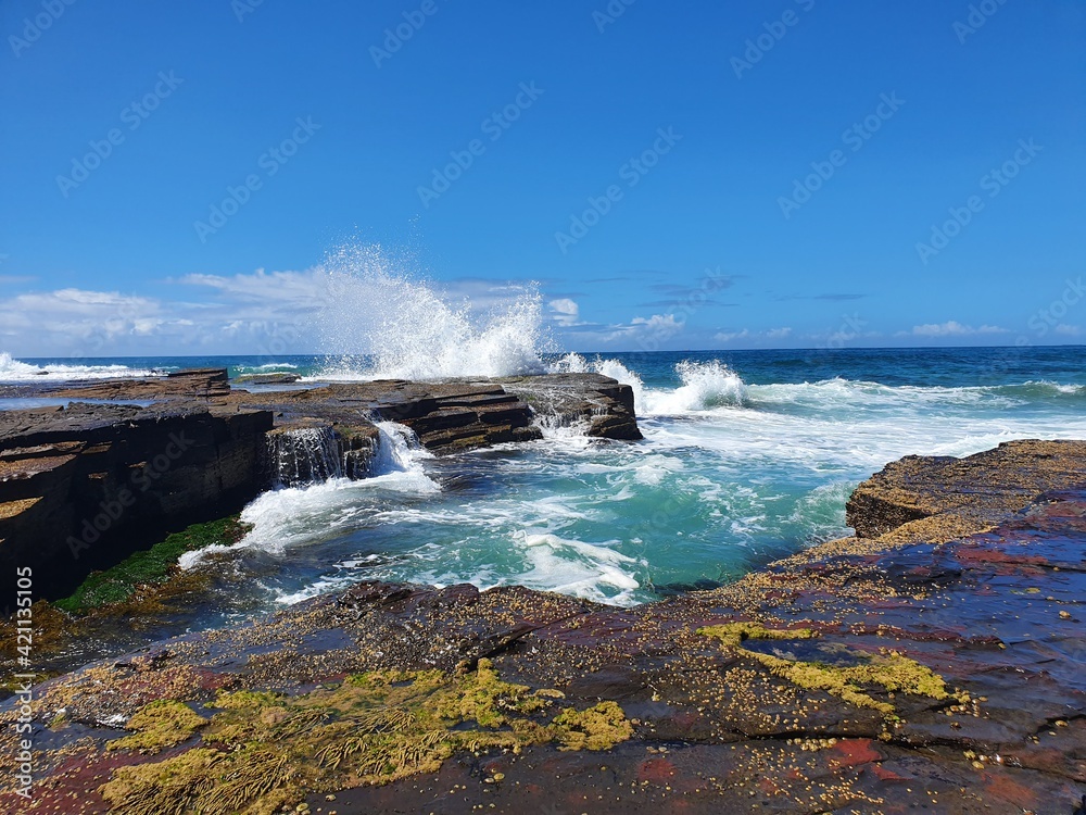 Waves crashing over coastal rocks on sunny blue sky day.