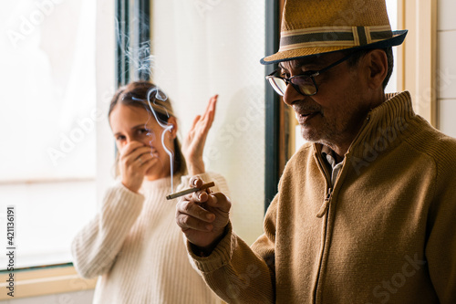 Elderly man smoking a cigarette with a young girl in the background bothered by breathing in the smoke that it gives off.