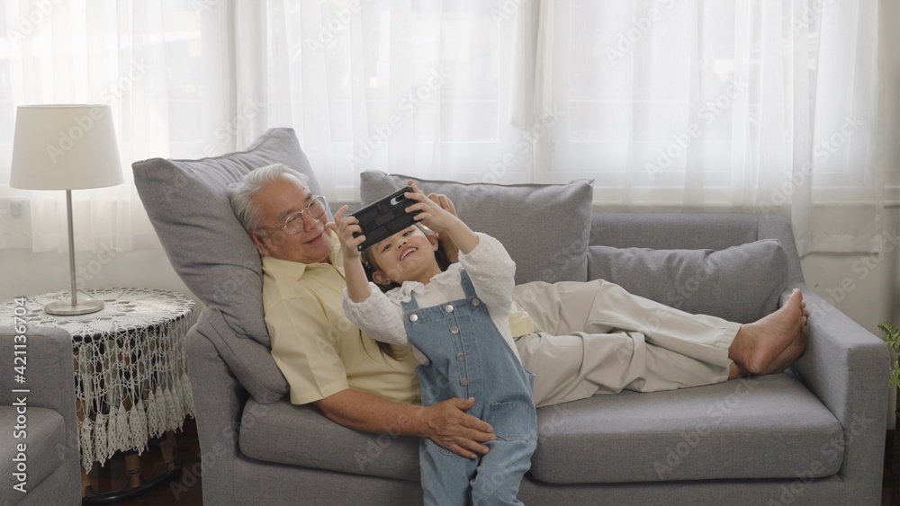 Portrait shot of the happy smiles Asian grandfather and little granddaughter taking selfie photos while sitting on the couch at home. Happy family with spending time together in living room.