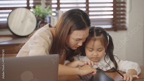 Young asian businesswoman mother using the laptop for working online from home with cute little girl drawing on tablet in the morning during quarantine of global coronavirus epidemic.