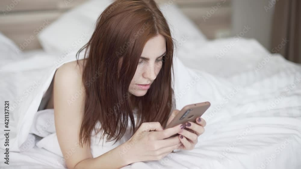 beautiful red-haired woman lying on bed at home during quarantine. Woman chatting using smartphone beautiful red-haired woman lying on bed at home during quarantine. Woman chatting using smartphone