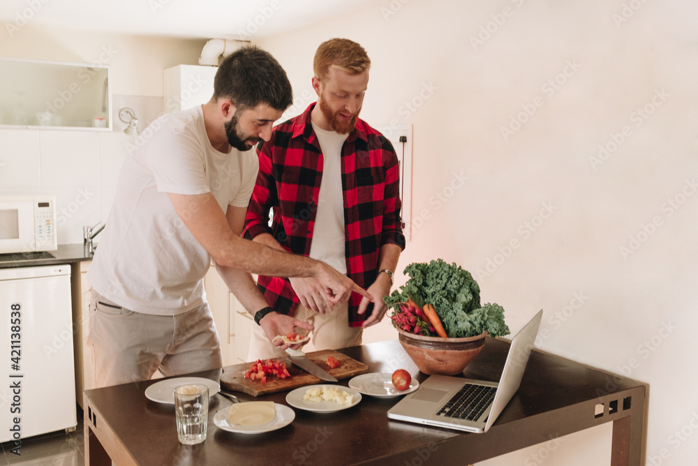 Young Male Couple Cooking While Looking At The Online Recipe On Their Laptop Young Male Couple Cooking While Looking At The Online Recipe On Their Laptop