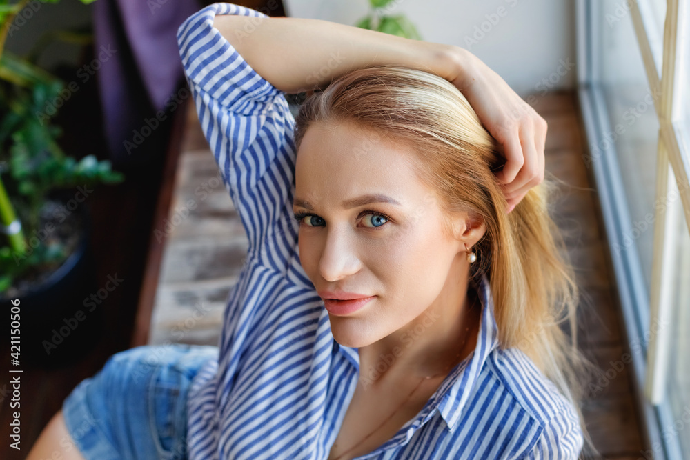 Fototapeta premium A young woman sits in front of the window on a large wooden sill. The girl is sitting on the windowsill. A young blonde is sitting by the window.