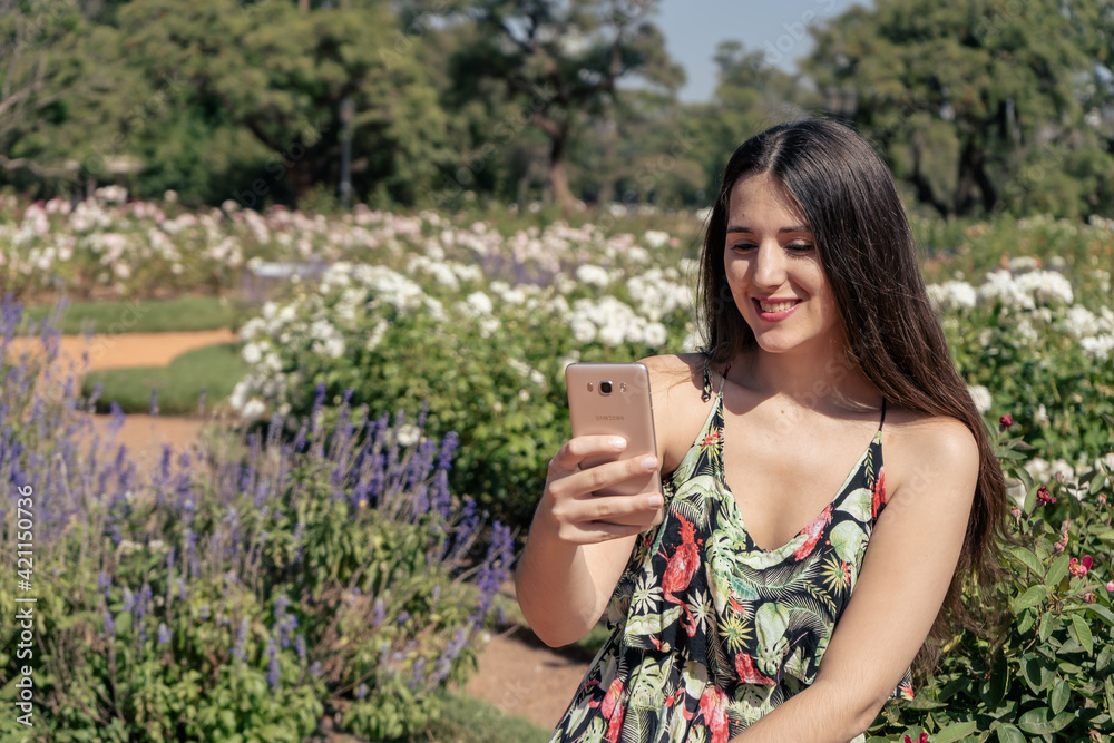Beautiful young Latin woman in a park receiving text messages on her cell phone. Technology and communication concept.