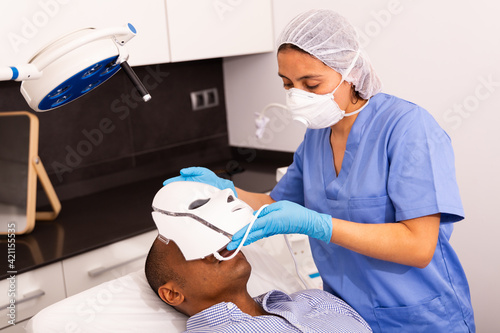 Hispanic female beautician preparing male client for procedure of phototherapy  putting LED mask on mans face