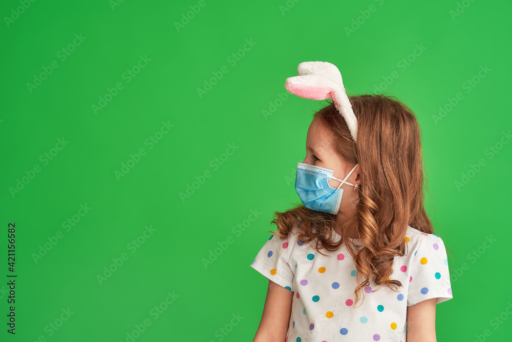 cute little girl wearing Easter bunny ears and mask standing on green background on Easter day. child is happy and celebrates Easter exactly according to the new rules. Protection from coronaviruses cute little girl wearing Easter bunny ears and mask standing on green background on Easter day. child is happy and celebrates Easter exactly according to the new rules. Protection from coronaviruses