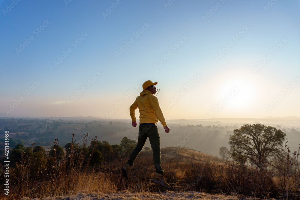 Young Mexican male wearing a yellow hoodie and cap jumping up in a field against a sunset sky