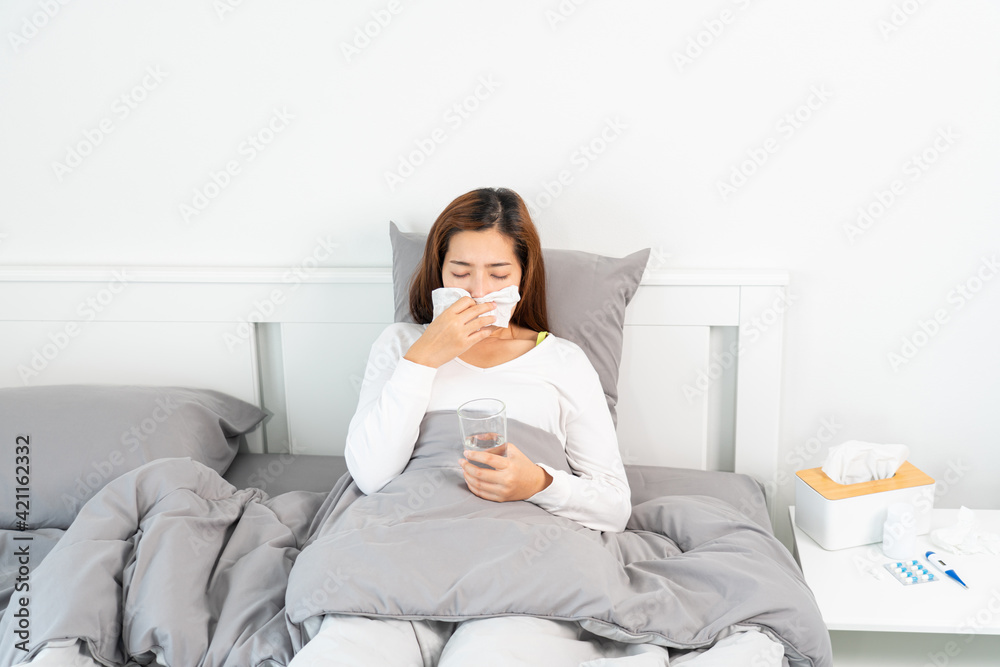 Young Asian Woman with seasonal infections Cold Blowing Her Nose and sneezing into Tissue with headache lying under the blanket in bed with high fever and a flu.