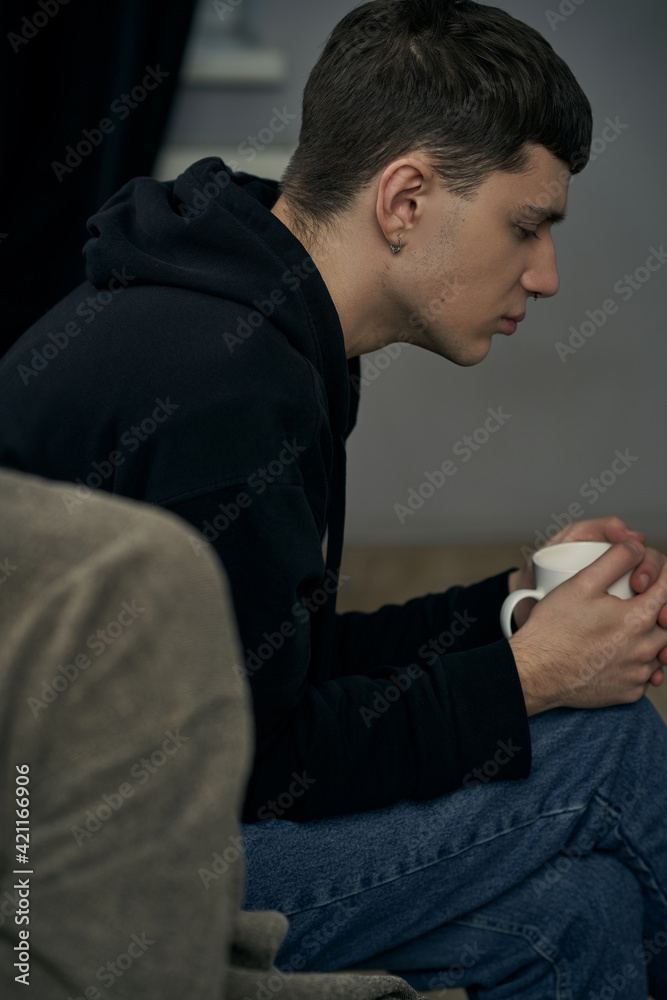 Staged photo illustrates problems and conflicts in gay couple relationships. Moment of showdown: upset black-haired man is sitting near his partner and holding white cup. 