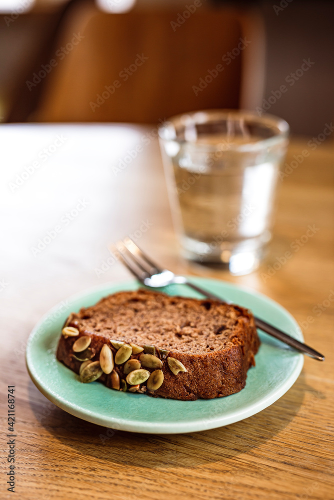 A slice of banana bread with pumpkin seeds on a blue plate, a dessert fork, a glass of water. Wooden table, cafe. A slice of banana bread with pumpkin seeds on a blue plate, a dessert fork, a glass of water. Wooden table, cafe.