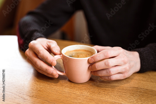 Man holding a pink ceramic cup of hot black coffee in his hands, sitting at the wooden table in a cafe.