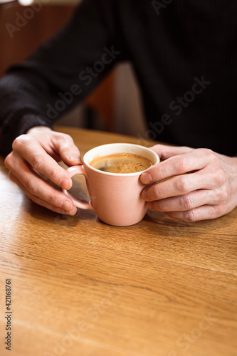 Man holding a pink ceramic cup of hot black coffee in his hands  sitting at the wooden table in a cafe. Copy space.