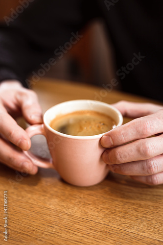 Man holding a pink ceramic cup of hot black coffee in his hands, sitting at the wooden table in a cafe. Close-up photo