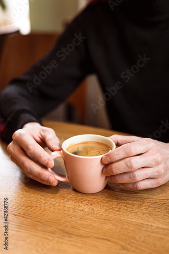 Man holding a pink ceramic cup of hot black coffee in his hands, sitting at the wooden table in a cafe.
