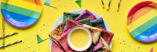 Rainbow party, table setup. Vibrant crepes on plate with honey. Party table, yellow paper tablecloth, drink straws, flags, rainbow paper plates. Rainbow as symbol of inclusivity, LGBT pride. photo