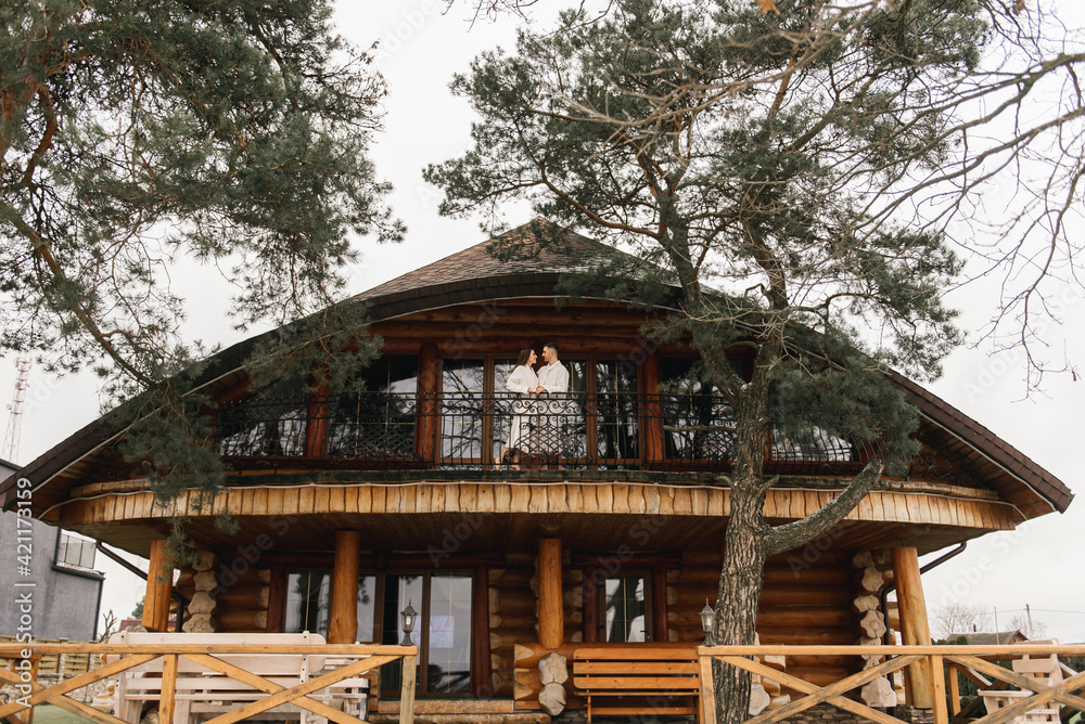 Young beautiful happy newlyweds in love a man and a woman in white bath robes on the balcony of the house smile and hug